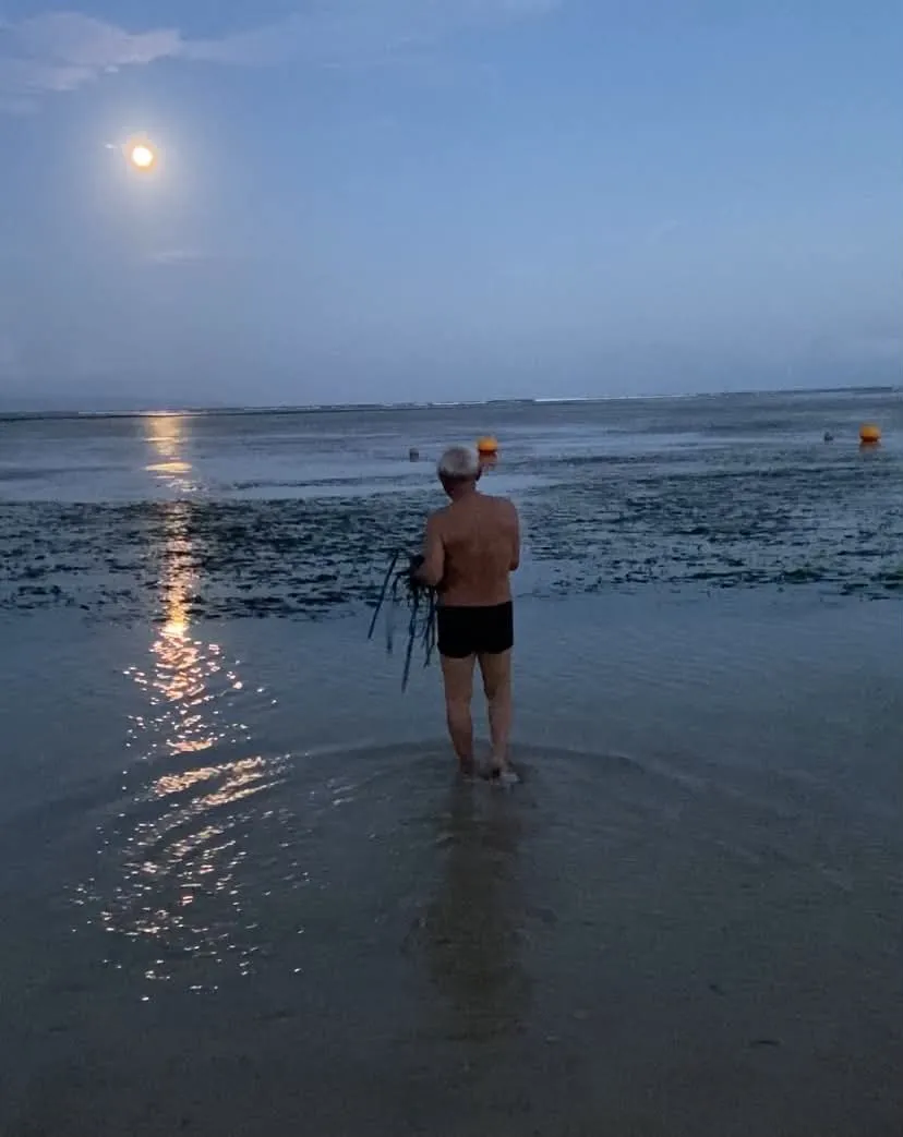 Muž stojící v mělké vodě během večerního odlivu za úplňku na pobřeží v Sanuru na Bali.