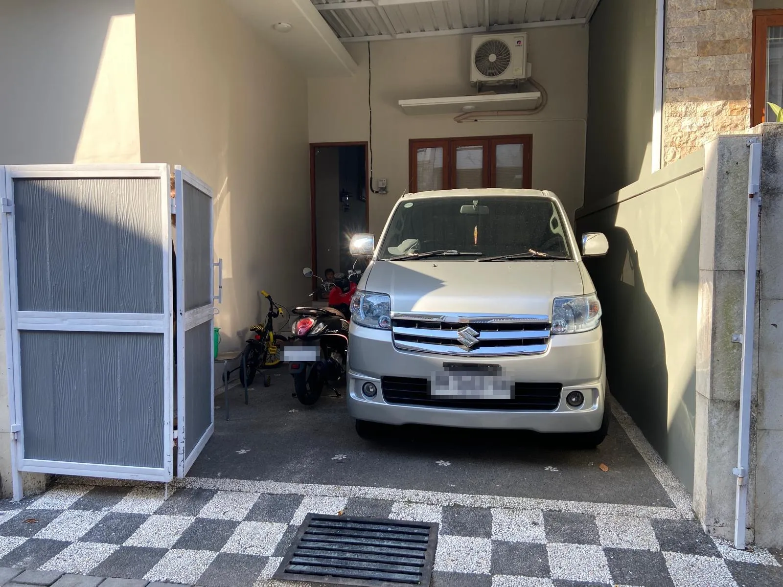 Auto a skútr zaparkované u domu na Bali, nad nimi venkovní jednotka klimatizace